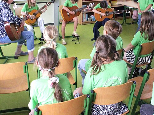 Die Kinder sitzen im Klassenraum der Trinitatisschule. Einige sitzen vor der Tafel und haben Gitarren in der Hand. Sie probieren die Instrumente aus.