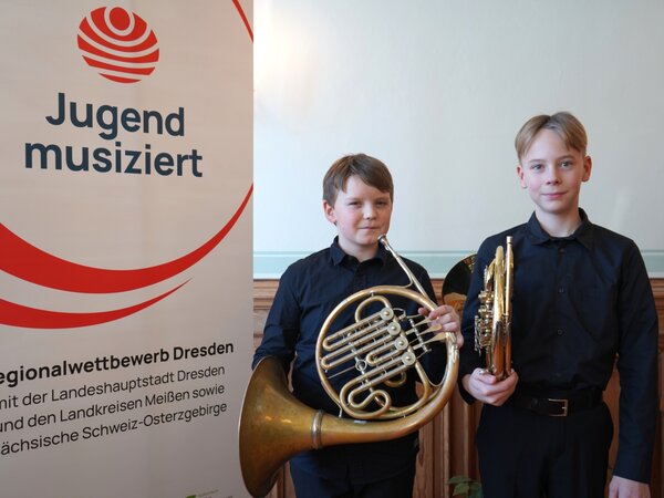 Zwei Hornschüler stehen neben dem Banner von Jugend musiziert und schauen in die Kamera. Sie halten stolz ihre Instrumente.