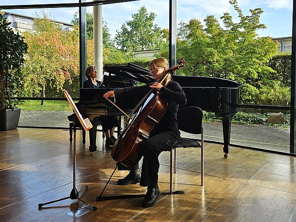 Eine Celloschülerin wird beim musizieren von einer Lehrerin am Klavier begleitet. Im Hintergrund sieht man Bäume und Büsche durch die großen Fenster. 