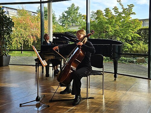 Eine Celloschülerin wird beim musizieren von einer Lehrerin am Klavier begleitet. Im Hintergrund sieht man Bäume und Büsche durch die großen Fenster. 