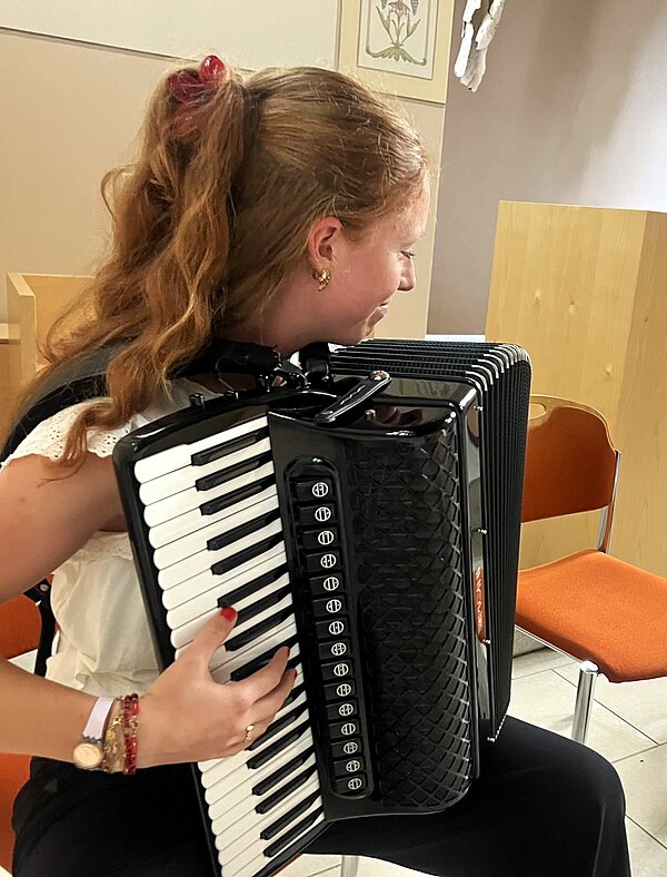 Eine Person mit zusammengebundenen, welligen Haaren spielt ein schwarzes Akkordeon mit Tasten und Knöpfen. Im Hintergrund stehen ein Stuhl und gerahmte Bilder an einer hellen Wand.