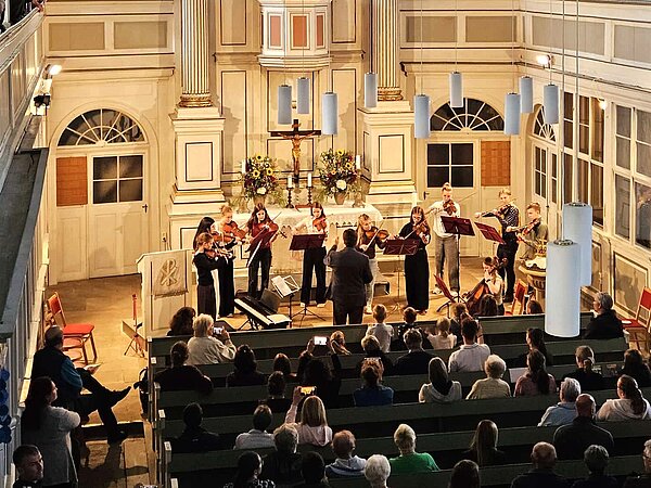 In einer Kirche spielt ein kleines Streichensemble vor dem Altarbereich. Eine dirigierende Person steht davor und leitet die Musikerinnen und Musiker an. Das Publikum sitzt in den Kirchenbänken.