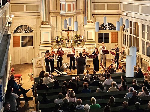 In einer Kirche spielt ein kleines Streichensemble vor dem Altarbereich. Eine dirigierende Person steht davor und leitet die Musikerinnen und Musiker an. Das Publikum sitzt in den Kirchenbänken.