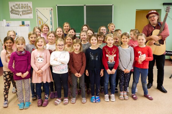 Die Kinder des Chores der Grundschule Niederau und Gerhard Schöne mit seiner Gitarre lachen in die Kamera.