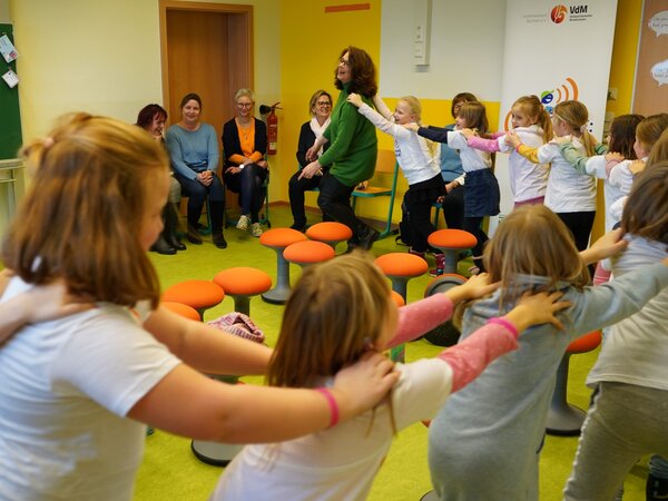 Die Kinder tanzen eine Polonaise durch das Klassenzimmer. Die Chorlehrerin führt den Zug an. Im Hintergrund sieht man Publikum in der Ecke sitzen.