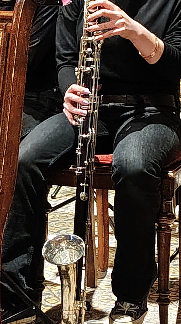 Auf dem Foto sitzt eine Person in dunkler Kleidung auf einem Holzstuhl und spielt eine Bassklarinette. Das große Instrument mit seinem gebogenen Metalltrichter ruht auf dem Boden, während die Hände des Spielers die Klappen greifen.