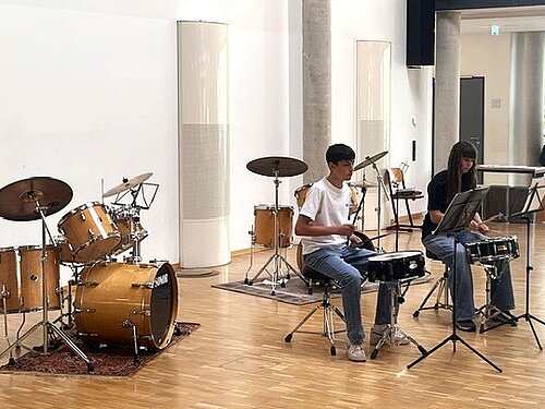 Zwei Personen sitzen in einem hellen Musikraum und üben an einzelnen Snare Drums. Vor ihnen stehen Notenständer, im Hintergrund sind zwei komplette Schlagzeuge aufgebaut.
