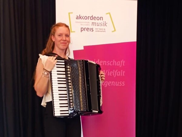 Eine Akkordeonschülerin präsentiert sich mit ihrem Akkordeon vor dem Banner des Akkordeon Musik Preises.
