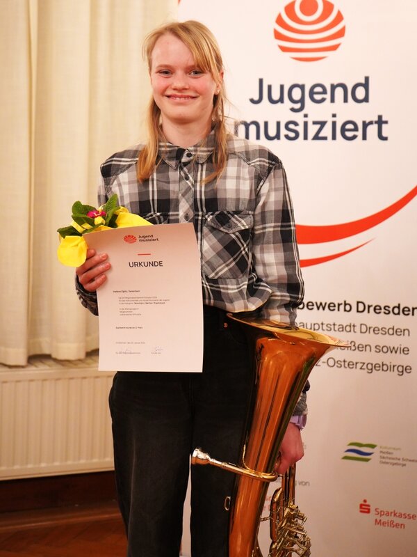 Eine Tubaschülerin steht vor dem Banner von Jugend musiziert und lacht in die Kamera. Sie hat ihre Urkunde und einen Blumentopf in der Hand. Die Tuba in der anderen Hand.