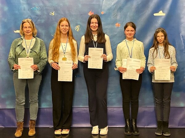 Vier Akkordeonschülerinnen und Ihre Lehrerin stehen vor einer blau-lila Wand. Sie haben Medaillen um den Hals und zeigen lachend ihre Urkunden in die Kamera.