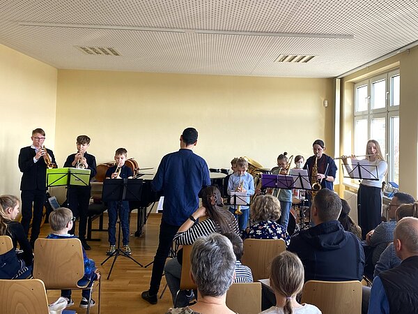 Schüler mit Blechblas- und Holzblasinstrumenten spielen gemeinsam vor einem Publikum. Vor ihnen steht ein Lehrer der dirigiert.