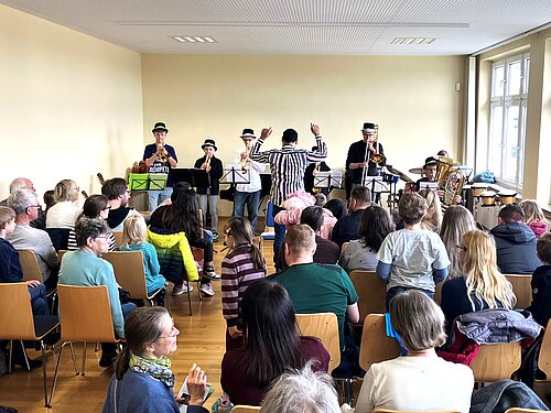 Zu sehen ist ein lebendiges Konzert in einem freundlich gestalteten Raum der Musikschule. Eine Gruppe von Musikerinnen und Musikern – ausgestattet mit Trompeten, Posaune, Tuba, Schlagzeug und Gitarre – spielt gemeinsam vor einem aufmerksam zuhörenden Publikum.