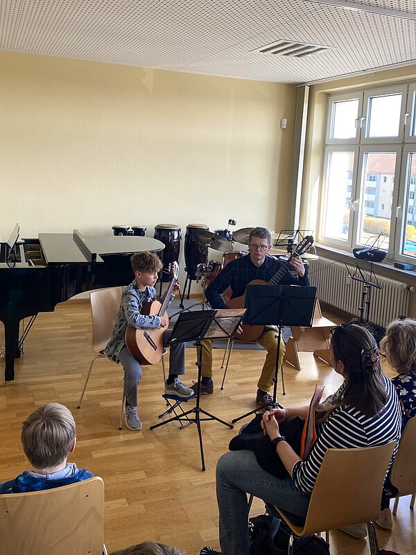Ein Gitarrenlehrer und sein Schüler spielen gemeinsam vor einem Publikum.