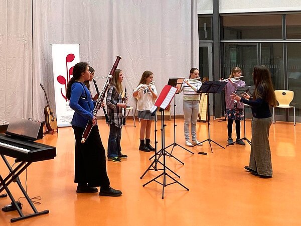 Ein Holzblasensemle spielt und wird dirigiert von einer Lehrerin. 