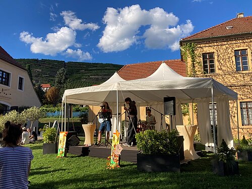 Eine junge Band steht auf der Bühne unter einem Zeltpavillion und spielt vor Publikum. Im Hintergrund sieht man das Weingut und die Weinberge.