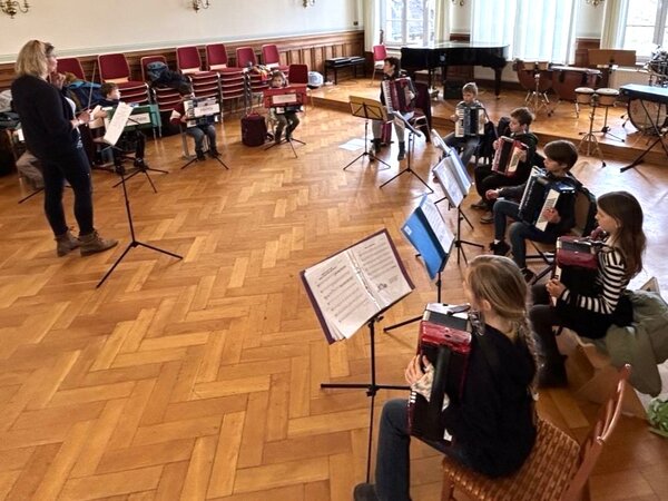 Es sitzen rechts in einem großen Saal Akkordeonschüler und musizieren zusammen. Ihre Lehrerin steh links im Bildund dirigiert.