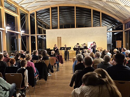 In einem modernen, hellen Saal mit hoher, gewölbter Decke und großen Glasfenstern stehen fünf Musikerinnen und Musiker an der Front des Raumes. Sie spielen Blechblasinstrumente, darunter Trompeten und eine Tuba, und stehen hinter Notenständern. Vor ihnen sitzt ein Publikum verschiedener Altersgruppen in mehreren Reihen und schaut aufmerksam zu.