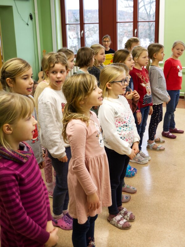 Ein Kinderchor steht in einem Klassenzimmer und singt. Im Hintergrund sieht man die Chorlehrerin am E-Piano spielen.