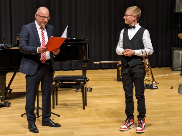 Max steht auf der Bühne, hat seinen Arm durch die Trompete geschoben und hält sie so fest. Er schaut zur linken Seite und hört freudig dem Moderator zu.
