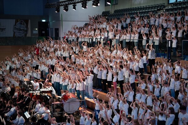 Viele Kinder stehen auf der Tribüne und haben die Arme und Hände in die Luft gestreckt. Alle tragen ein weißes T-shirt mit dem Logo von "Stimme an".