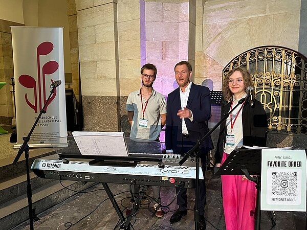 Nathalie und Moritz von der Coswiger Band "Favorite Order" stehen zusammen mit Ministerpräsident Michael Kretschmer in der sächsischen Staatskanzlei. Es ist außerdem das Logo der Musikschule und ein Keyboard vor dem goldenen Tor zu sehen.