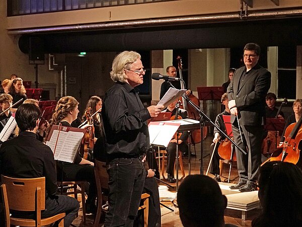 Der bekannte deutsche Schauspieler Thomas Stecher steht am Mikrofon und rezitiert Texte. Hinter ihm sitzt das Jugendsinfonieorchester Radebeul und Matthias Herbig und verfolgt seine Worte.