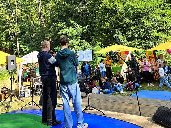 Zwei Musikschüler stehen auf einer Bühne im Wald mit ihren Trompeten und musizieren. Das Publikum sitzt unter Sonnenschirmen und auf Decken und hört aufmerksam zu.