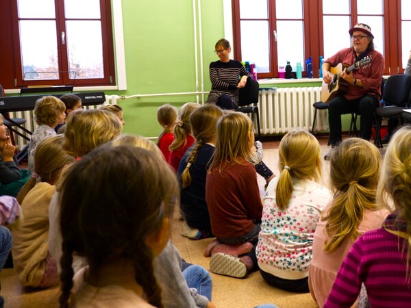 Kinder sitzen auf dem Fußboden in einem Klassenzimmer. Sie hören Gerhard Schöne beim Singen und Gitarre spielen zu. Links hinter den Kindern steht ein E-Piano.  An der Wand auf einem Stuhl sitzt lachend die Chorlehrerin.