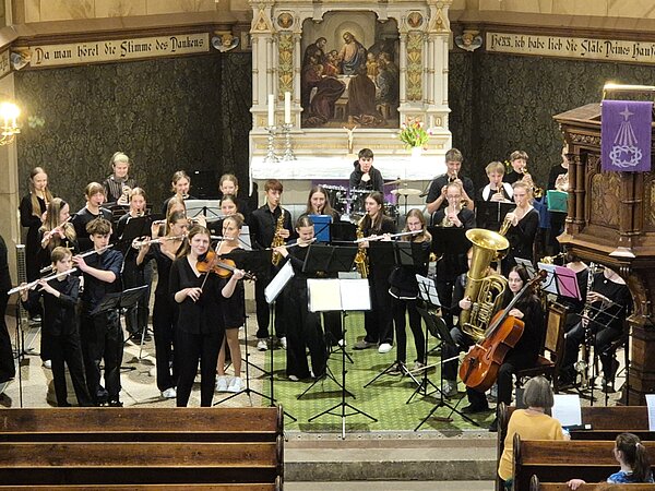 Das Foto zeigt ein Jugendensemble, das in einer Kirche musiziert. Die jungen Musikerinnen und Musiker sitzen in mehreren Reihen und spielen verschiedene Instrumente, darunter Flöten, Violinen, Saxofone, Trompeten, Tuba, Cello und Schlagwerk. Alle tragen schwarze Konzertkleidung. Im Hintergrund ist der reich verzierte Altarraum mit religiösen Kunstwerken und Kerzen zu sehen. An der Kanzel hängt ein violettes Tuch mit christlichem Symbol. 