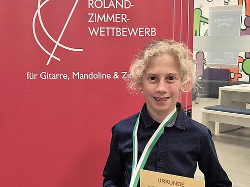 Ein Junge lächelt in die Kamera. Hinter ihm sieht man das Banner vom Roland-Zimmer-Wettbewerb.