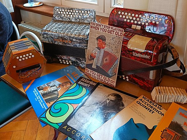 Auf einem Tisch stehen zwei große Akkordeons – eines grau, eines rot – sowie ein kleineres Instrument. Davor liegen mehrere Notenhefte und Schallplatten zum Thema Akkordeon.