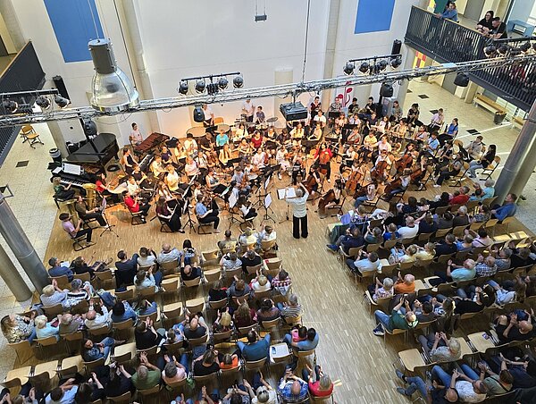 Ein großes Orchester spielt vor Publikum. Die Ansicht ist aus der Vogelperpesktive.