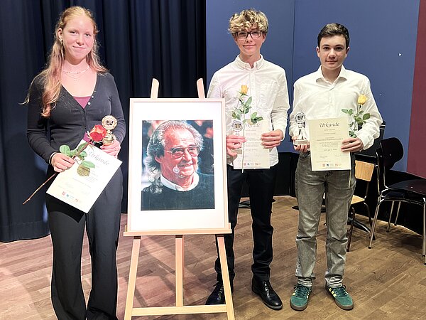 Schüler stehen und schauen freundlich in die Kamera. Sie halten in den Händen Urkunden und Blumen. Dazwischen steht ein Fotoportrait von Werner Richter.