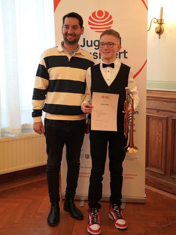 Ein Schüler und sein Lehrer stehen vor dem Banner von Jugend musiziert und strahlen in die Kamera. Der Schüler hält seine Urkunde in der einen und seine Trompete in der anderen Hand.