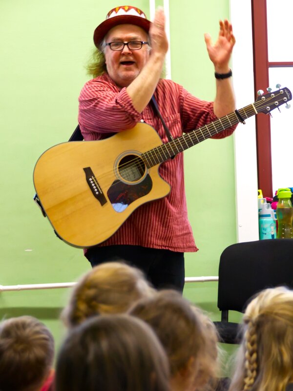 Gerhard Schöne steht vor den Kindern. Er hat seine Gitarre um und klatscht rechts oben in die Hände.