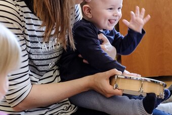 Ein Kleinkind schlägt freudig auf eine Trommel während es auf dem Schoß seiner Mutter sitzt.