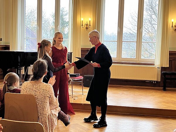 Zwei Schülerinnen bekommen ihre Urkunden überreicht in einem festlichen Raum. Sie werden von von der Musikschulleiterin Frau Haas und dem Publikum mit Applaus geehrt.