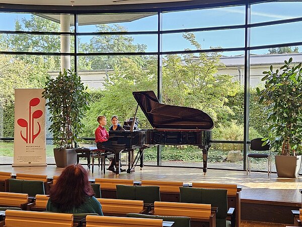 Junger Pianist spielt am Flügel, während seine Lehrerin neben ihm sitzt; im Vordergrund sitzt Publikum im Saal der Landesärztekammer Sachsen.