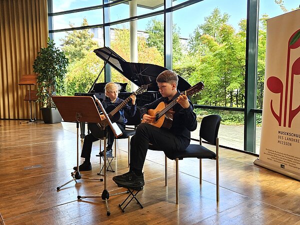 Zwei Gitarrenschüler spielen zusammen auf einer Bühne. Im Hintergrund steht ein Flügel und Büsche scheinen durch die Fenster.