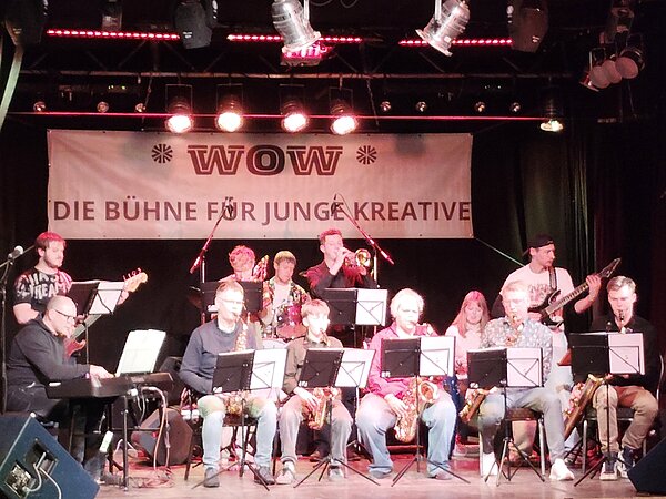 Auf einer Bühne sitzt und steht eine größere Gruppe junger Musikerinnen und Musiker in mehreren Reihen. Sie spielen verschiedene Instrumente, darunter Saxophone, Trompete, Posaune, E‑Gitarre, Bass, Keyboard und Schlagzeug. Vor ihnen stehen Notenständer. Über der Bühne hängt ein großes Banner mit der Aufschrift „WOW – Die Bühne für junge Kreative“. 