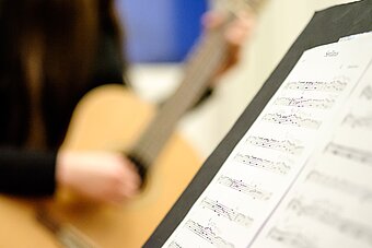 Im Vordergrund sind schräg Noten zu erkennen. Im Hintergrund verschwommen ein Gitarrenschüler.
