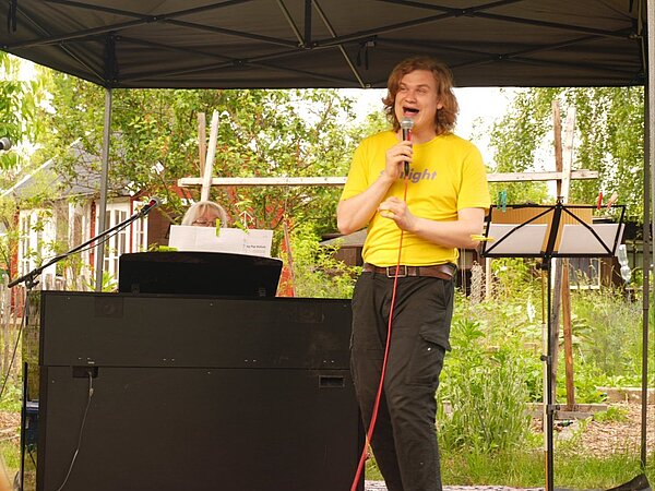 Wilhelm Dulig singt mit Freude und Mikrofon in der Hand. Er wird vom Klavier begleitet und steht unter einem Pavillon im Grünen.