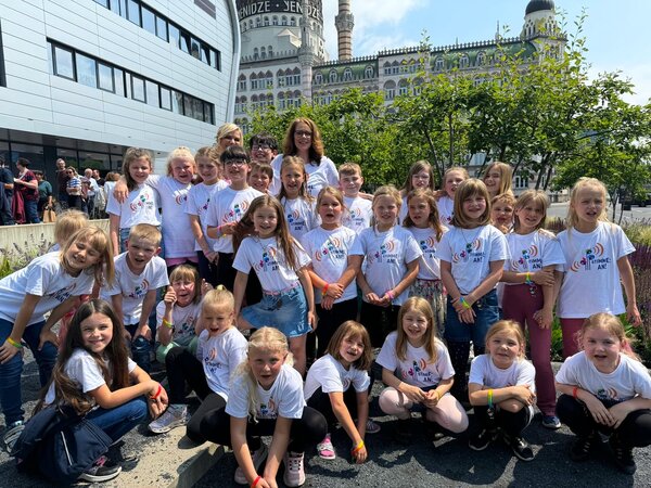 Die Kinder des Kinderchores der Grundschule Ponickau mit Ihrer Lehrerin Frau Taubert lächeln in die Kamera. Im Hintergrund sieht man Bäume, links die Ballsportarena und dahinter das Jenidze.