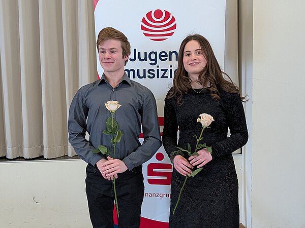 Auf dem Foto stehen zwei junge Musikerinnen bzw. Musiker nebeneinander vor einem Banner des Wettbewerbs „Jugend musiziert“. Beide halten eine weiße Rose in der Hand. Die Person links trägt ein graues Hemd und schwarze Hose, die Person rechts ein langes schwarzes Kleid.