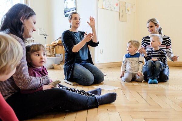Musikgartenkinder sitzen mit ihren Eltern im Kreis und schauen zur Lehrerin die mit ihren Händen ein Finderspiel zeigt.