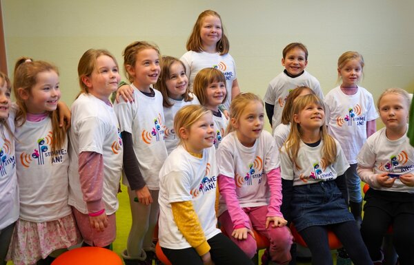 Die Kinder des Kinderchores der Grundschule Ponickau posieren für die Kamera. alle Kinder tragen ein weißes T-Shirt mit dem Logo von Stimme an.
