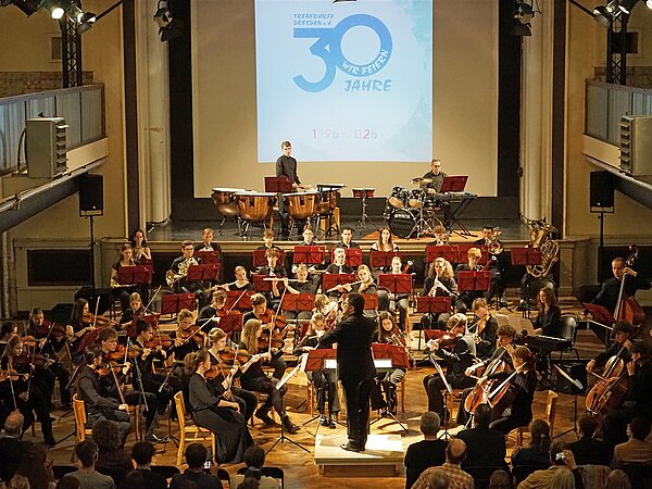 Jugendsinfonieorchester Radebeul musiziert im großen Saal des Zentralwerks Dresden unter Leitung von Matthias Herbig. Im Hintergrund ist ein Bild an der Leinwand zum Jubiläum von 30 Jahren Treberhilfe Dresden e.V. zu sehen.