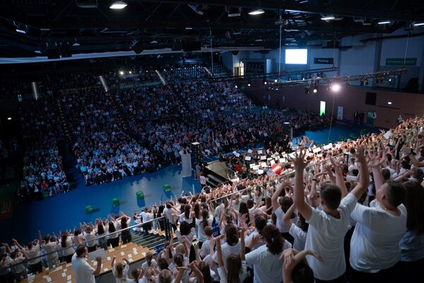 Viele Kinder stehen auf der Tribüne und haben die Arme und Hände in die Luft gestreckt. Gegenüber sitzt das zahlreiche Publikum.