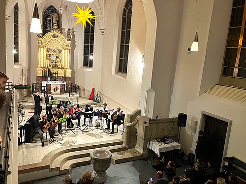 Ein Gitarrenensemble, welches von Bläsern begleitet wird, spielt vor Publikum in der Klosterkirche.