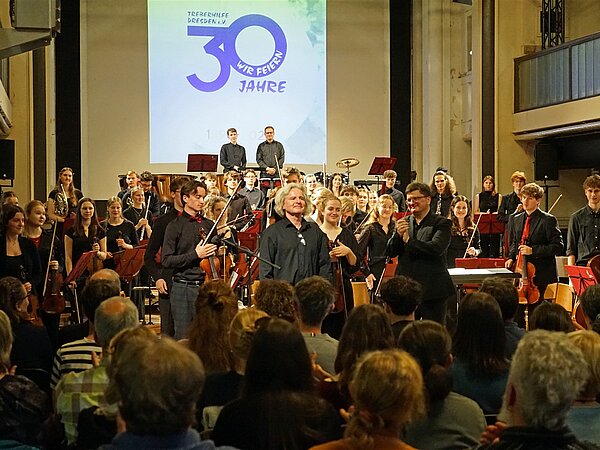 Der bekannte deutsche Schauspieler Thomas Stecher steht mit dem Jugendsinfonieorchester Radebeul und Matthias Herbig auf der Bühne des Zentralwerk Dresden. Im Hintergrund ist ein Bild an der Leinwand zum Jubiläum von 30 Jahren Treberhilfe Dresden e.V. zu sehen.